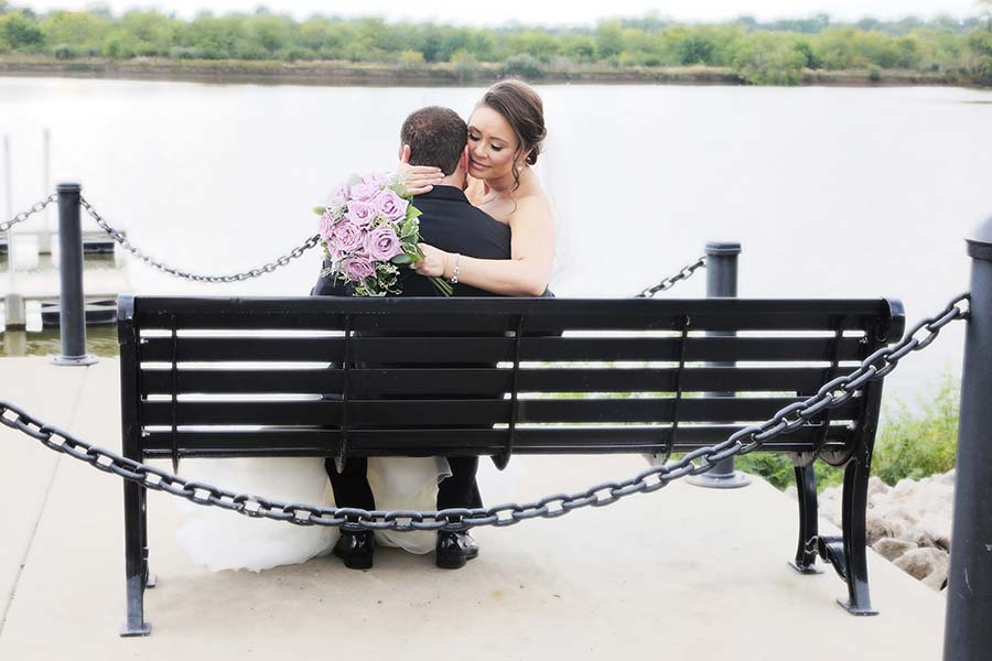 the bride is sitting on her new husband's lap on a park bench on the Maumee River in downtown Perrysburg before the reception at Stone Oak country club in Holland Ohio