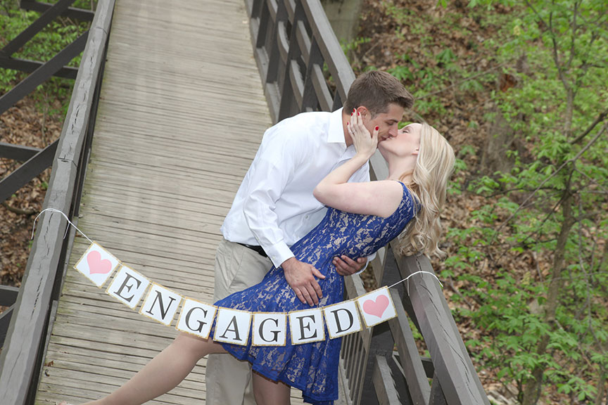the groom is kissing the bride while he is dipping her on the wooden bridge with an engaged sign in front of them