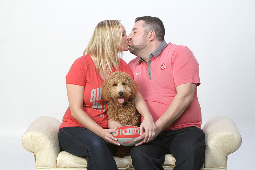 engaged couple wearing ohio state shirts and sitting on a cream settee with their dog in between them at Kurt Nielsen Photography Portrait Studio in Sylvania, Ohio