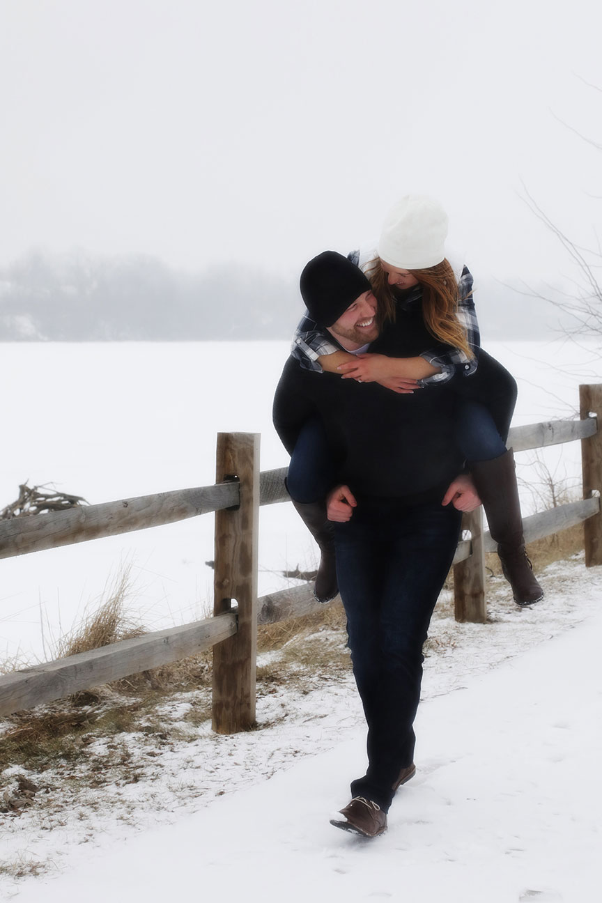 man and woman laughing as the man gives the woman a piggy back ride while walking along the boardwalk near the maumee river in downtown Toledo in the winter with snow on the ground