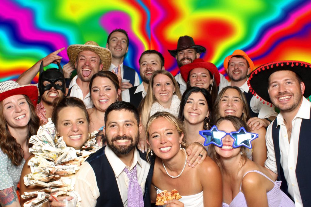 bride and groom surrounded by their friends who were wearing props like hats and oversized glasses and featherless boas while posing for a photo booth photo