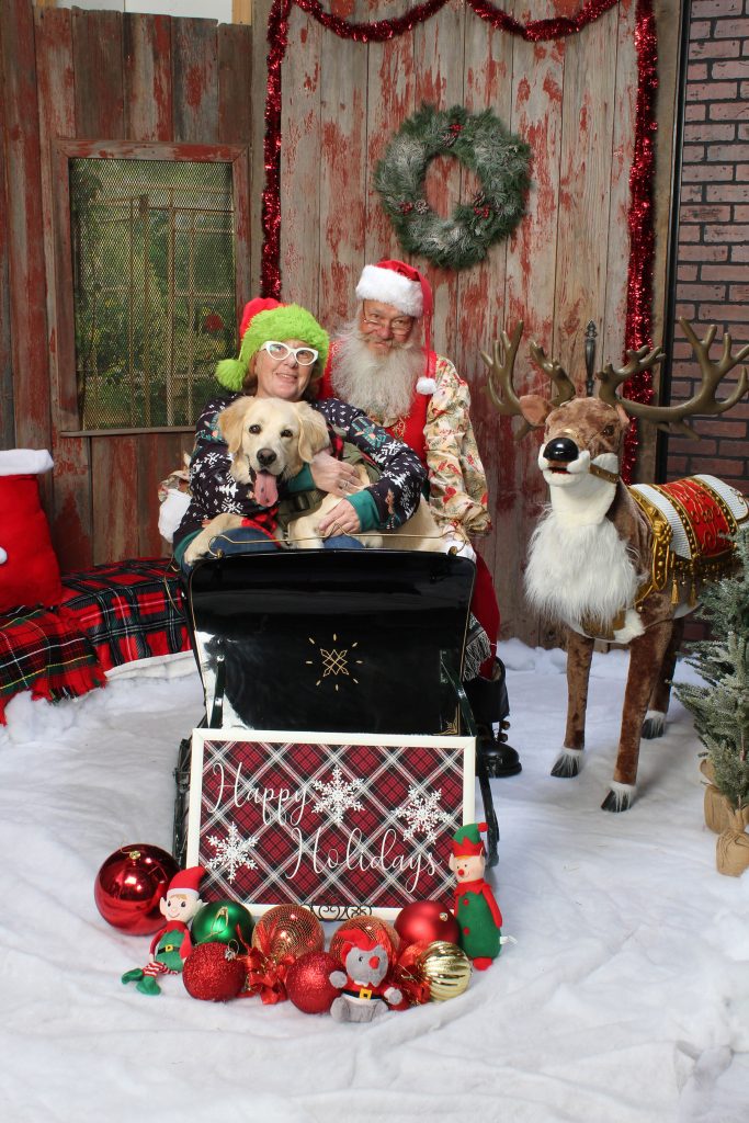 mom and her golden retriever dog sitting in Santa's sleigh with Santa behind them in the barn set at Kurt Nielsen Photography