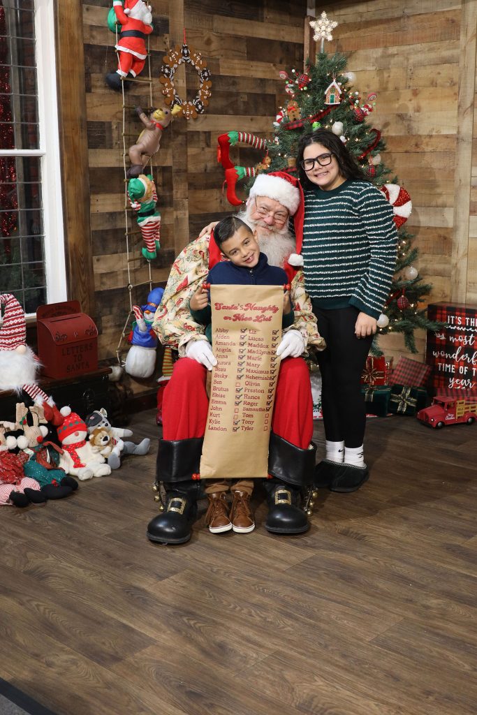 a little boy is sitting on Santa's lap while holding Santa's Naughty and Nice List with his big sister standing beside them inside Kurt Nielsen Photograpy photo studio